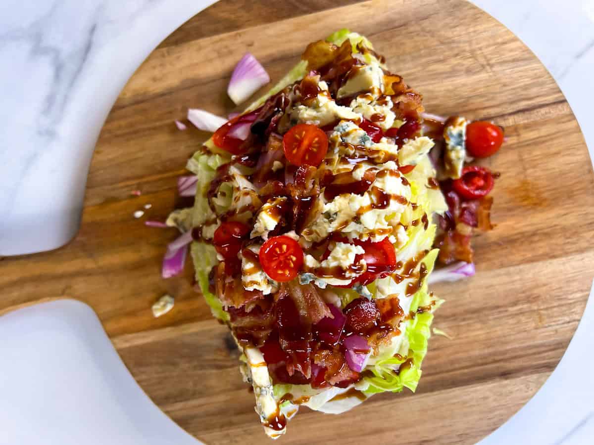 Finished blue cheese wedge salad with toppings on a round wooden cutting board.