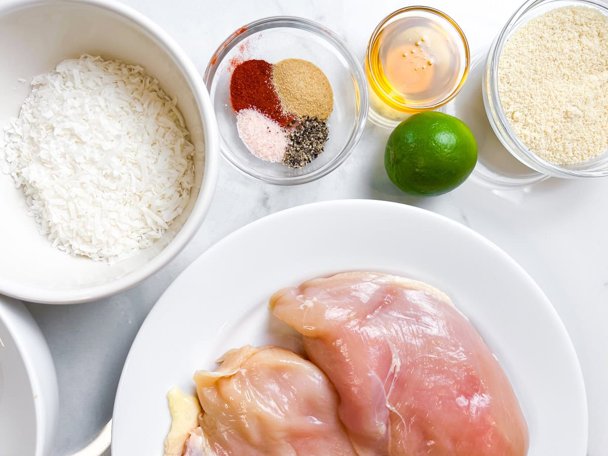 Raw chicken, spices, almond flour, honey, and spices in bowls on a marble counter.