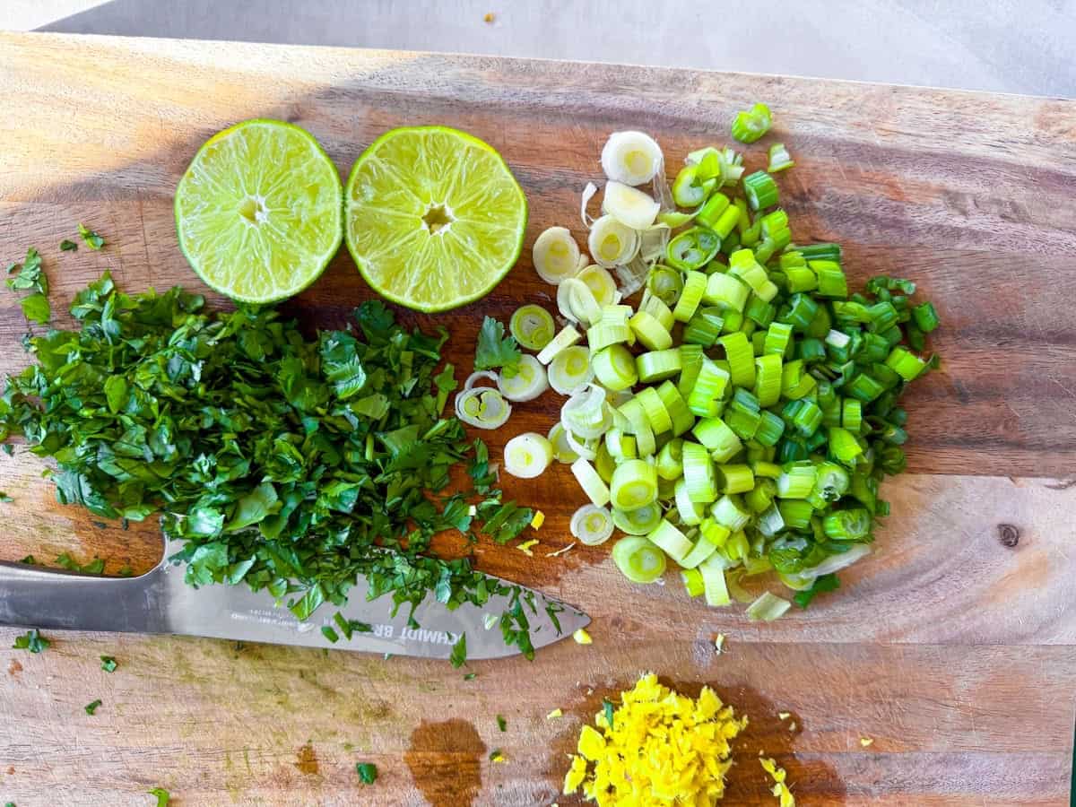 Chopped cilantro, green onion, minced ginger on a wooden cutting board with a knife.