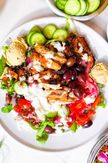 Cava style bowl with hummus, tzatziki, lentil tabbouleh, tomatoes, and pickled onions on a white plate.