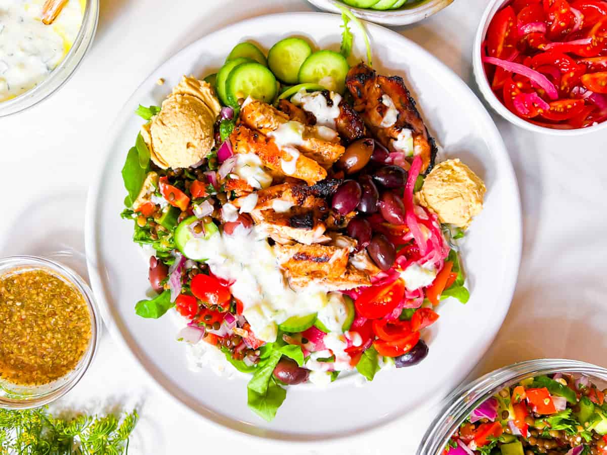 A Cava style bowl with chicken, rice, cucumbers, hummus, tzatziki, pickled onions, and cucumbers on a white plate.