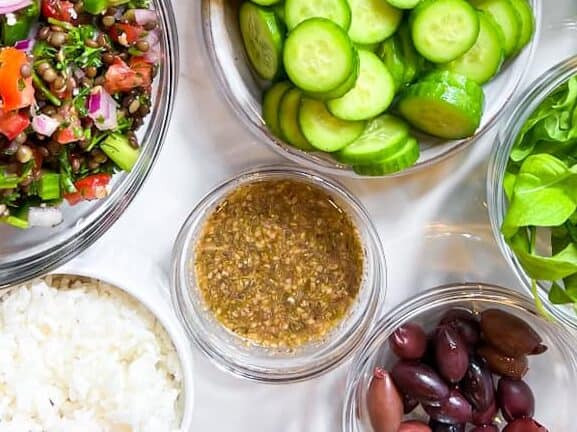 a clear bowl with a dark vinaigrette inside of it and other ingredients like cucumber and olives in bowls around it. 