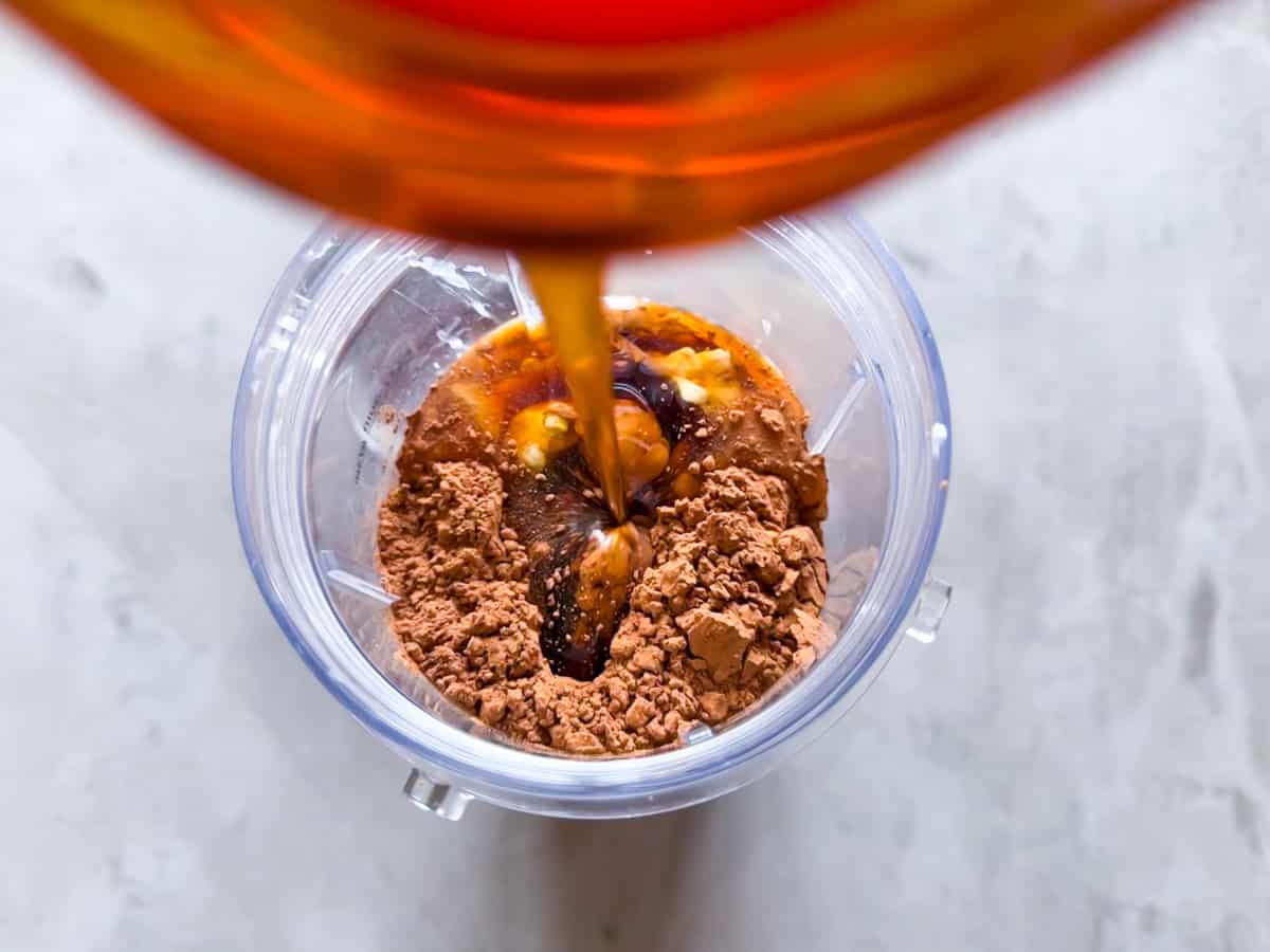 Maple syrup being poured into a blender with cocoa powder and cottage cheese in a blender cup.